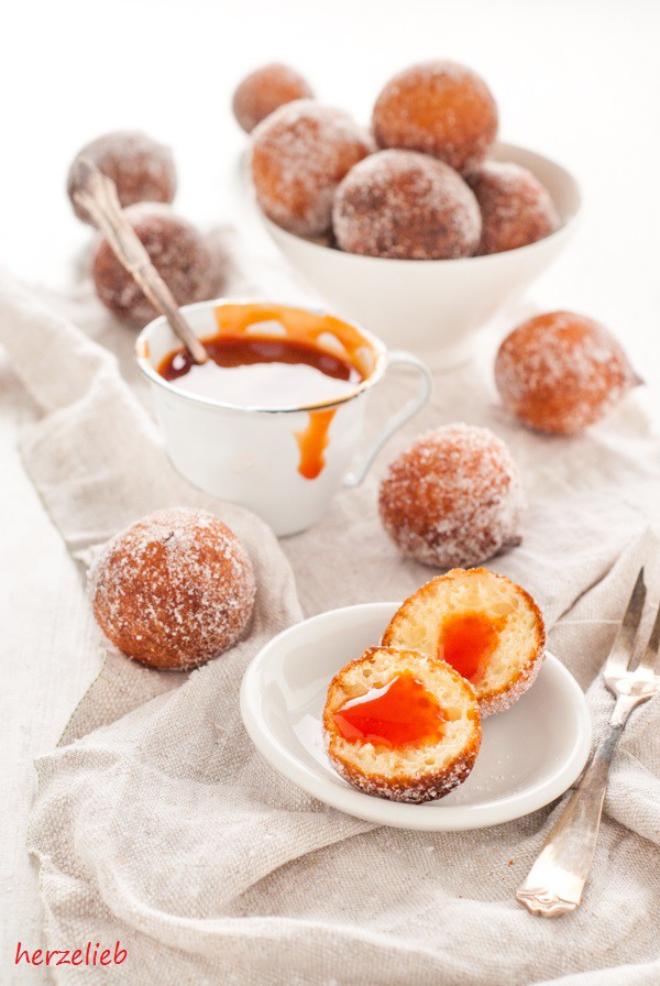 Auf diesem Bild zum Quarkbällchen Rezept von herzelieb sieht man im Vordergrund ein halbiertes Quarkbällchen mit Marmelade auf einem weißen Teller, daneben liegt eine Kuchengabel. Im Hintergrund weitere Bällchen, die mit Zucker bestreut sind. Weiter hinten eine Schale mit mehr Kuchen und einer Tasse mit Marmelade. 