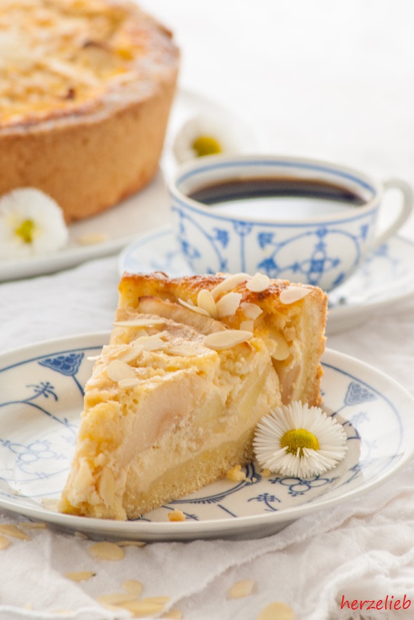 Ein Stück Apfelkuchen sieht man auf diesem Bild zum Apfel-Schmandkuchen Rezept. Der Schmandkuchen liegt auf einem weißblauem Teller. Dahinter rechts eine Tasse Kaffee, links der ganze Kuchen.