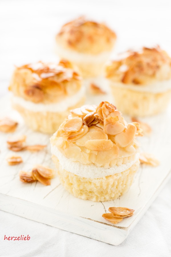 Auf diesem Bild zum Bienenstich Muffins Rezept sieht man 4 Bienenstichmuffins mit einer Füllung aus Creme und einem Topping aus karamellisierten Mandeln auf einem weißen Holzbrett. Dekoriert mit Mandelblättchen