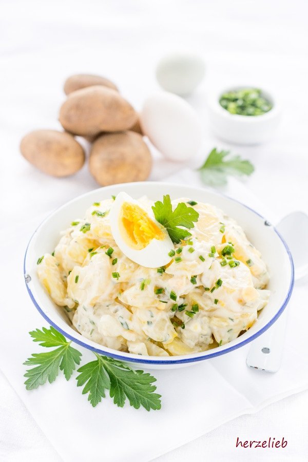 Grillsalat oder Partysalat in einer weißen Emaille Schüssel mit blauem Rand nach einem Kartoffelsalat Rezept mit Mayonnaise wie von Mutti. Dekoriert mit Ei, Schnittlauch und glatter Petersilie. Im Hintergrund: Kartoffeln, zwei weiße Eier, ein Schälchen mit Schnattlauchringen und ein Petersilienblatt. Neben der Salatschüssel liegt ein weiße Löffel aus Emaille.