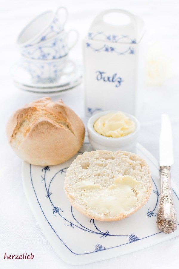 Dieses Bild zum Rundstykker Rezept zeigt ein halbes dänisches Brötchen, mit Butter bestrichene. Dahinter ein Schälchen mit Butter und ein ganzes Brötchen. Dahinter ein Salzbehälter und 3 Tassen und Untertassen von einem blauweißen Geschirr.