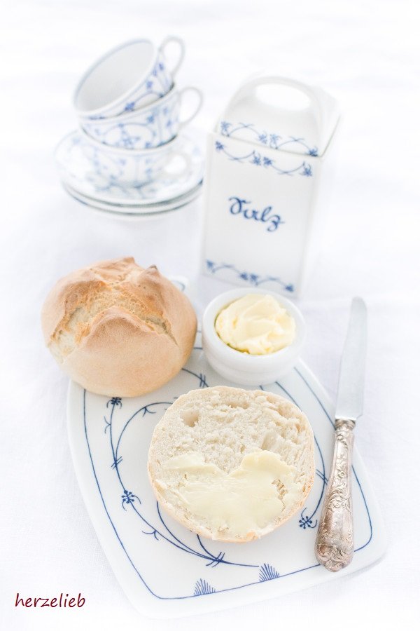 Dieses Bild zum Rundstykker Rezept zeigt ein halbes dänisches Brötchen, mit Butter bestrichen. Rechts daneben liegt ein silbernes Dessert. Dahinter ein Schälchen mit Butter und ein ganzes Brötchen. Dahinter 3 Tassen und Untertassen von einem blauweißen Geschirr.