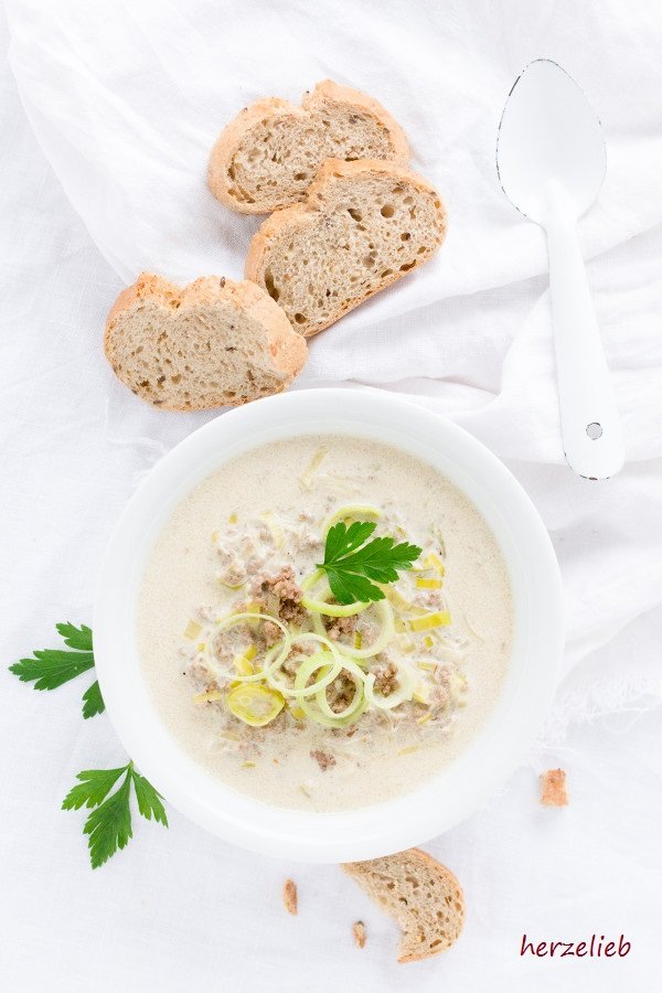 Dieses Bild zum Käse-Lauchsuppe mit Hackfleisch Rezept ist von oben fotografiert. In der Mitte der weiße Suppenteller mit der Suppe. Zu sehen ist die Suppe, in der Hackfleisch ist. Dekoriert mit Lauchringen und Petersilie. Im Vordergrund ein angebissenes Stück Brot, links 2 Blättchen Petersilie. Im Hintergrund ein weißer Emaillelöffel und 3 Stücke Brot