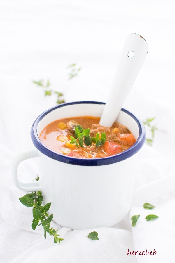 Einen Becher mit der Hackfleischsuppe sieht man auf diesem Bild zum Pizzasuppe Rezept. Die Partysuppe ist dekoriert mit Oregano und im Becher steckt ein Löffel. Dekoriert ist das Bild mit einem Kräuterzweig und Blättern.