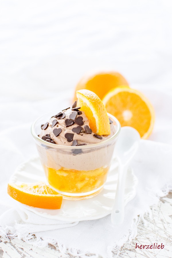 Ein Gläschen mit Schokoladen-Mousse mit Orangenkompott sieht man auf diesem Bild zum Dessert im Glas Rezept. Ganz unten eine leuchtend orangefarbene Schicht mit Orangenkompott, darüber die zartblauen Schokoladenmousse. Dekoriert mit Schokoladenherzen. Das Glas steht auf einem weißen Unterteller, im Vordergrund liegt ein Orangenschnitz. Hinter dem Nachtisch liegen eine ganze und eine halbe Orange.