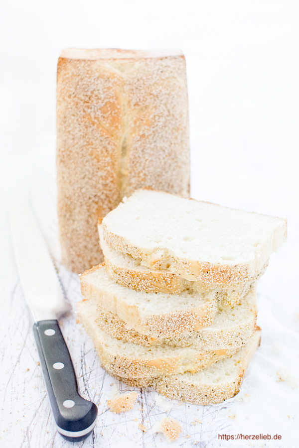 Auf diesem Bild zum Dänisches Franskbrød Rezept sieht man ein Weißbrot, dass aufrecht auf der Schnittfläche steht. Davor liegt ein Stapel mit 6 Brotscheiben. Links daneben liegt ein Brotmesser. 