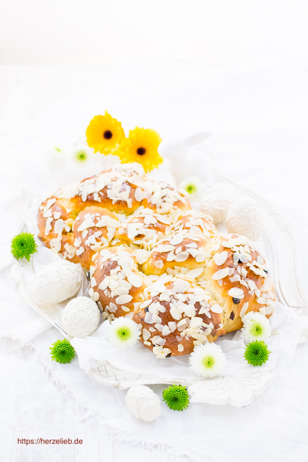 Auf diesem Bild zum Osterzopf Rezept sieht man das geflochtenen Brot mit Rosinen. Es ist mit Mandelblättchen bestreut und rundherum mit weißen und grünen Blüten dekoriert. Im Hintergrund einige gelbe Blüten.