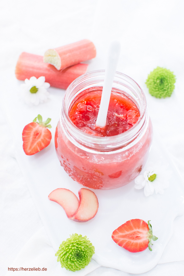 Das Foto zum Rhabarbermarmelade Rezept zeigt ein Glas Marmelade mit einem weißen Porzellanlöffel darin von oben fotografiert. Dekoriert mit halben Erdbeeren, Rhabarberscheiben und 2 Rhabarberstangen sowie grünen Blüten und einer Margerite im Hintergrund.