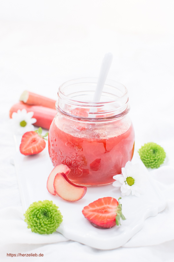 Auf diesem Bild zum Rhabarbermarmelade Rezept sieht man ein Glas mit Marmelade und einem Löffel darin. Es steht auf einem weißen Porzellanbrett. Dekoriert mit weißen Blumen, grünen Blüten, halben Erdbeeren, Rhabarberscheiben und zwei Rhabarberstangen im Hintergrund.