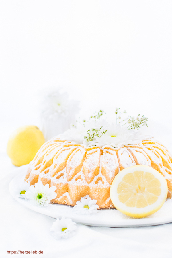 Auf diesem Bild zum Zitronenkuchen Rezept sieht man den ganzen Grießkuchen Gugelhupf auf einem weißen Teller. Dekorier mit Zitronen, weißen Blüten und Blumen in der Mitte. 