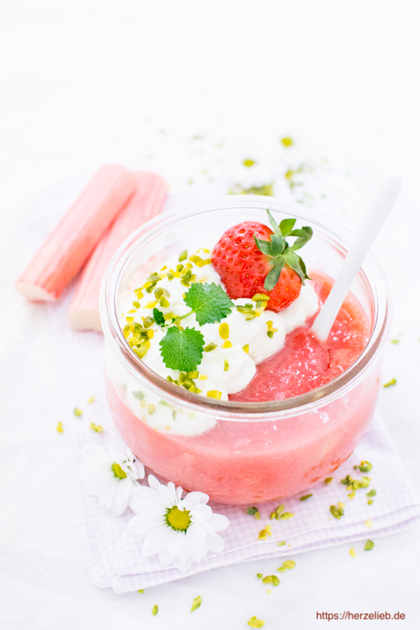 Dieses Foto zum Rhabarber Dessert Rezept zeigt die Rhabarbergrütze im Weckglas mit Erdbeeren, Sahne und Pistazien. Dekoriert mit Margeritenblüten, Rhabarberstangen, gehackter Pistazie, Zitronenmelisse Blätter und einer frischen Erdbeere.
