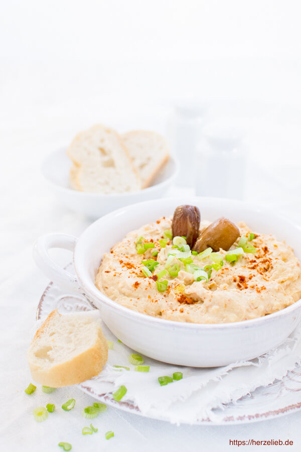 In einer weißen Schale ist ein Dattel Dip mit Harissa, Datteln und Frühlingszwiebel-Ringen angerichtet.Im Vordergrund eine Brotscheibe. Im Hintergrund Gewürzstreuer und eine Schale mit Brot. 