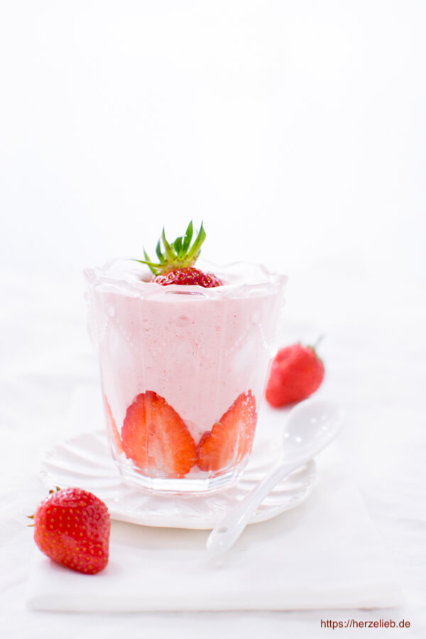 Auf diesem Foto zum Erdbeer Dessert Rezept sieht man das Erdbeerdessert im Glas sieht man ein Glas mit dem Nachtisch. Auf einer Erdbeerschicht am Boden kommt ein rosafarbenes Mousse und obenauf eine halbe Erdbeerscheibe. Dekoriert mit ganzen Erdbeeren.
