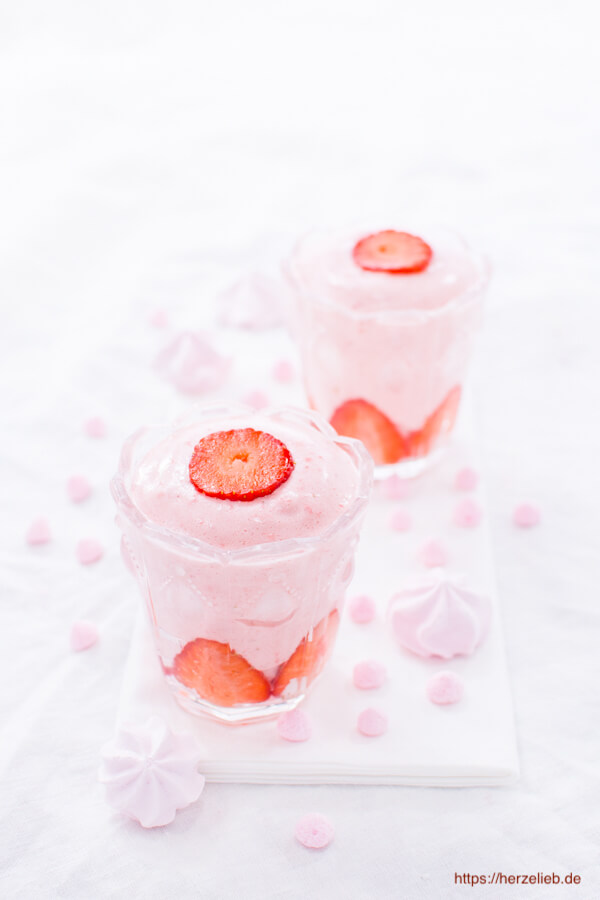 Auf diesem Bild zum Erdbeerdessert Rezept sieht man das Erdbeer Dessert im Glas sieht man zwei Gläser mit dem Nachtisch. Auf einer Erdbeerschicht am Boden kommt ein rosafarbenes Mousse und obenauf eine Erdbeerscheibe. Dekoriert mit Rosa Baiser Tuffs.