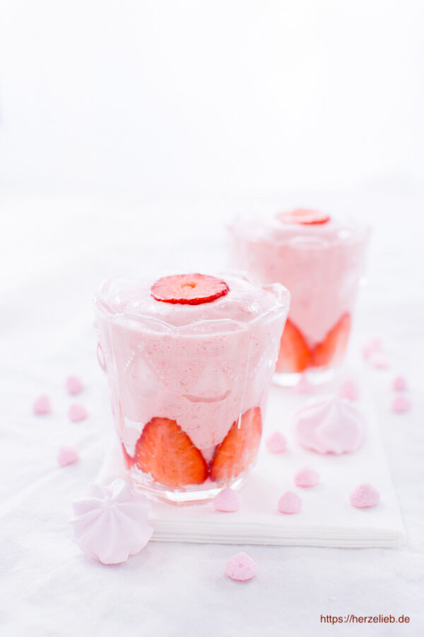 Auf diesem Bild zum Erdbeer Dessert Rezept sieht man 2 Gläser mit dem Nachtisch Dekoriert mit rosa Baiser. Auf einer Erdbeerschicht am Boden kommt ein rosafarbenes Mousse und obenauf eine halbe Erdbeerscheibe. Dekoriert mit ganzen Erdbeeren.