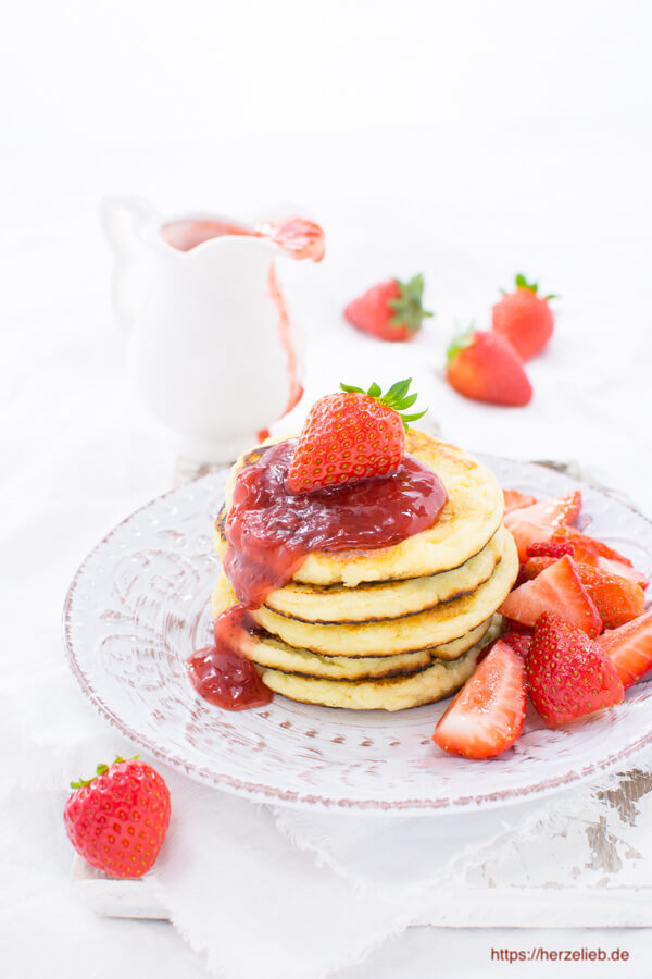 Ein Stapel Grießbreipfannkuchen sieht man auf diesem Bild Grießbrei Pfannkuchen Rezept. Angerichtet mit roter Fruchtsoße und frischen Früchten bzw. Erdbeeren. Im Hintergrund liegen 3 Erdbeeren und daneben steht ein Kännchen mit roter Fruchtsauce.