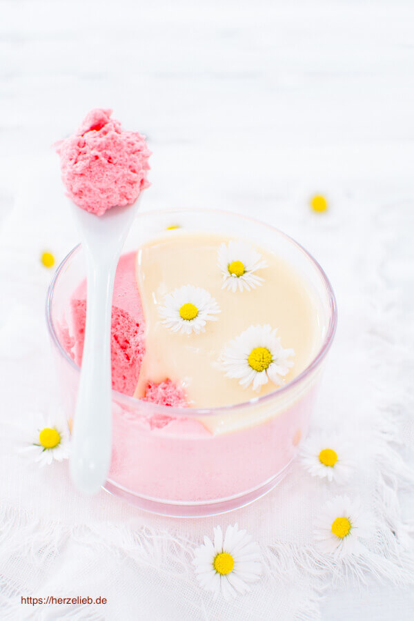 Auf diesem Bild zum Mädchenröte Dessert im Glas Rezept ist ein Glas mit dem Nachtisch zu sehen. Das rosafarbene Dessert ist mit Vanillesauce begossen. Dekoriert mit Gänseblümchen. Auf dem Dessert ein Löffel mit dem flufüigen Dessert. Dekoriert rundherum mit Blümchen