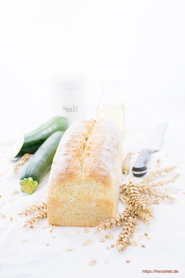 Dieses Foto zum Zucchinibrot zeigt ein ganzes Zucchinibrot. Im Hintergrund 2 Zucchini, ein Salzbehälter aus Porzellan und ein Brotmesser. Dekoriert mit Getreideähren.