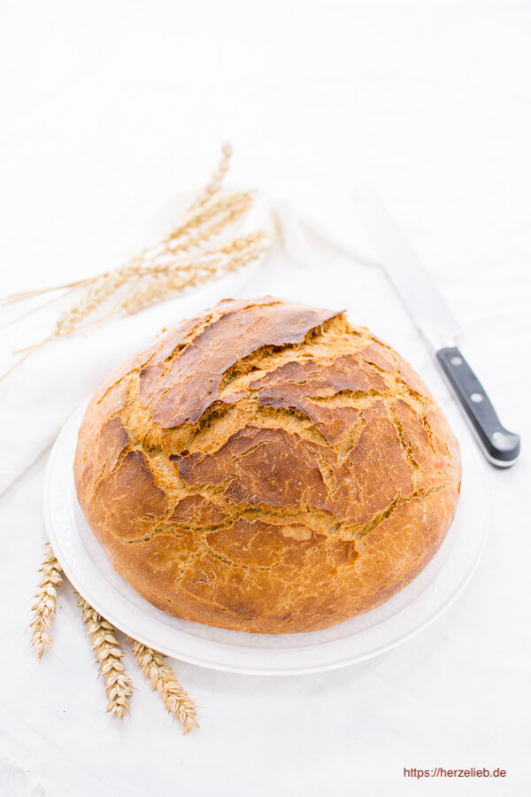 Auf diesem Bild zum Dinkel-Roggenbrot Rezept sieht man den ganzen goldbraun gebackenen Brotlaib auf einem weißen Teller. Dekoriert mit Getreideähre. Hinten rechts ein Brotmesser. 