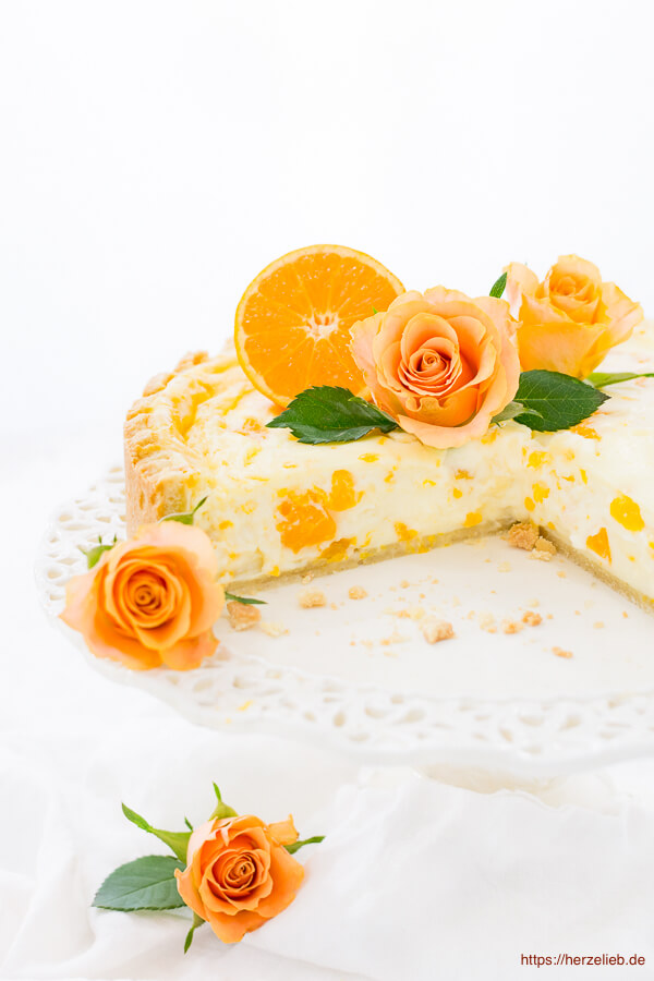 Einen angeschnitten Schmandkuchen sieht man auf diesem Bild zu Omas Mandarinen Schmand Kuchen Rezept mit einer Prise Liebe. Dekoriert ist die Schmandtorte mit orangefarbenen Rosen, Blättern vom Orangenbaum und einer halben Orangen. 