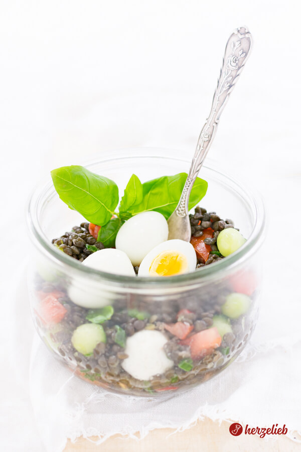 Dieses Bild für das Belugalinsen-Salat Rezept mit Ei, Mozzarella, Tomate, Gurke und Frühingszwiebeln ist von schräg oben fotografiert. Der Linsensalat im Glas ist dekoriert mit Basilikum, Wachteleiern, Gurkenkugeln und Tomatenswürfeln. im Salat steckt eine Gabel.