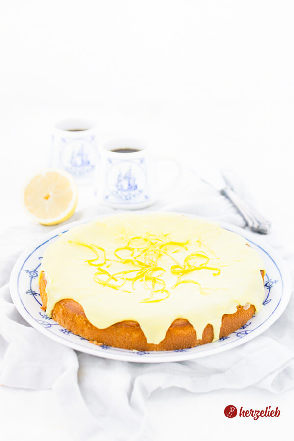 Auf diesem Bild zum Zitronenkuchen Rezept sieht man den selbstgebackenen Citronmåne aus Dänemark. Ein goldbraun gebackener Kuchen mit einer strahlend gelben Glasur. Dekoriert mit Zitronenzesten. Der Kuchen liegt auf einem blauweiße Teller. im Hintergrund Tassen mit einem Segelschiff und Kaffee darin, außerdem eine halbe Zitrone. Rechts hinten ein Tortenmesser.