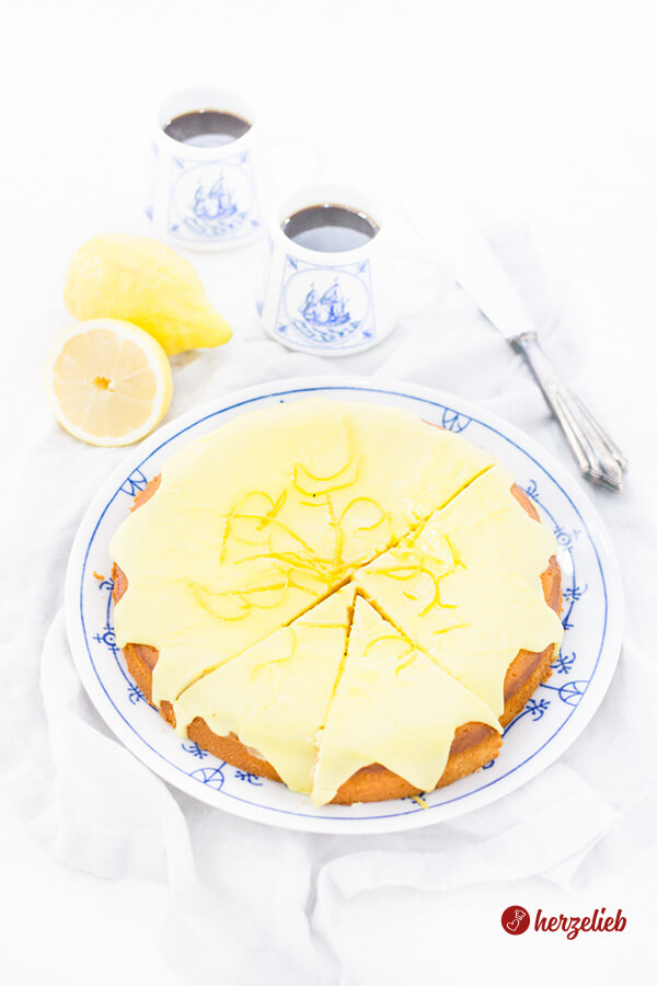Dieses Bild zum Citronmåne Rezept zeigt den dänischen Zitronenkuchen mit Marzipan auf einem blauweißen Teller und ist von oben fotografiert. Im Hintergrund sieht man zwei Becher mit Kaffee, eine halbe und eine ganze Zitrone und ein Kuchenmesser.