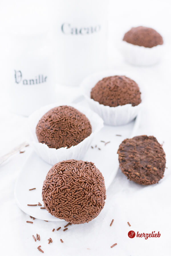 4 große Rumkugeln mit Schokoladenstreuseln in weißen Muffinförmchen sieht man auf diesem Bild zum Rumkugeln Rezept. Im Hintergrund ein Vanille- und ein Kakao Gefäß aus weißem Porzellan.