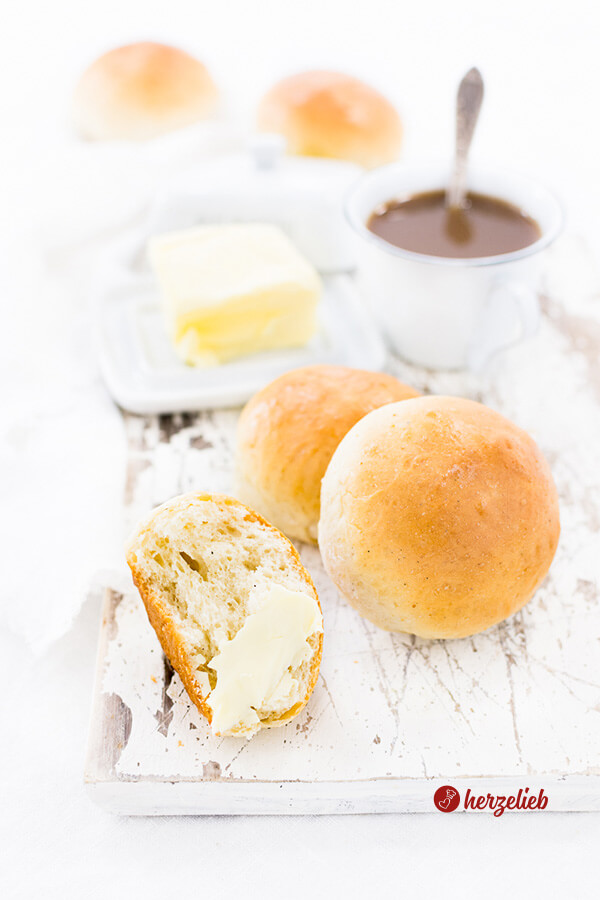 Auf diesem Bild zum Milchbrötchen Rezept sind 2 Brötchen mit einer Tasse Kaffee und einer Buttergkocke zu sehen. Dahinter zwei weitere Milchbrötchen. Im Vordergrund ein halbes Brötchen mit Butter bestrichen