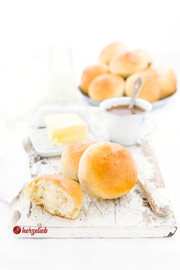 Auf diesem Bild zum Milchbrötchen Rezept sieht man im Vordergrund ein angebissenes Milchbrötchen. Dahinter zwei Brötchen die übereinander liegen. Seitlich ein Messer. Im Hintergrund eine Tasse Kaffe, eine geöffnete Butterglocke mit Butter und dahinter ein Korb voller  Milchbrötchen