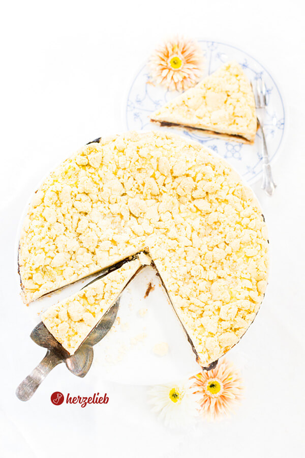 Dieses Foto zum friesischer Käsekuchen Rezept aus der Springform zeigt den Kuchen, der oben fotografiert ist. Der Kuchen ist angeschnitten, ein Stück liegt auf einem silbernen Kuchenhaber. Links oben im Hintergrund ein ein stück vom Streuselkuchen auf einem blauweißen Teller mit einer orangefarbenen Blüte und einer silbernen Kuchengabel. Im Vordergrund zwei Blüten. 