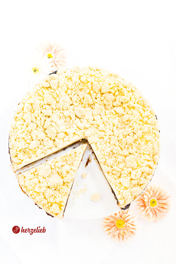 Dieses Bild zum friesischer Käsekuchen Rezept ist von oben fotografiert. Man sieht die Butterstreusel und das der Streuselkuchen mit Pflaumenmus gefüllt ist. Der Kuchen ist angeschnitten, ein Stück setzt ist hervorgezogen. Im Vordergrund 2 orangefarbene Blüten. Im Hintergrund zwei weitere Blüten.