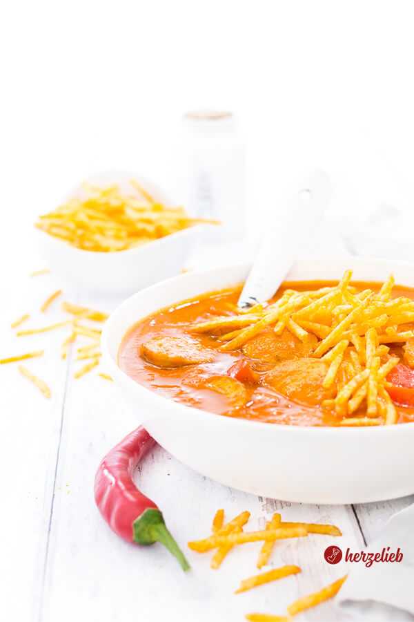 Dieses Bild zum Currywurstsuppe Rezept zeigt eine weiße Schüssel mit der Tomaten-Currysauce. In der Suppe sieht man Wurstscheiben, Paprikastücke und Kartoffelsticks. Im Hintergrund eine weiße Porzellanschale mit Kartoffel Sticks und ein Gewürzgefäß. Vorne ein rote Chilischote