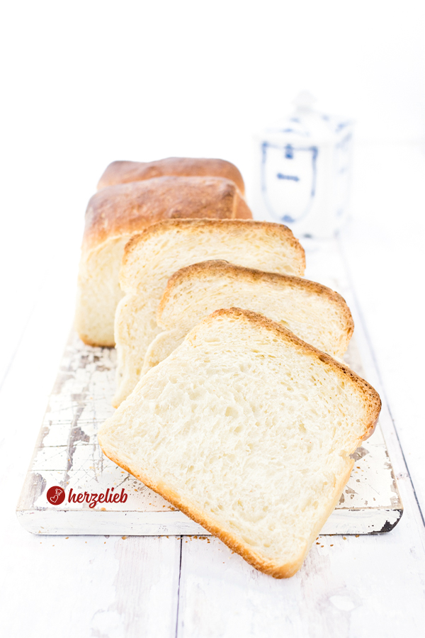 Auf diesem Bild zum Sandwichbrot Rezept sieht man das angeschnittene Brot. Im Vordergrund sind 3 abgeschnittene Scheiben. Das Brot liegt auf einem weißen Holzbrett. Im Hintergrund ist ein blau-weißer Honigtopf aus Porzellan zu sehen