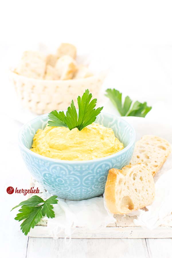 Auf diesem B ildCurry-Mango Dip Rezept vom Foodblog herzelieb sieht man eine blaue Schale mit dem gelben Currydip. Dekoriert mit glatter Petersilie, vor der Dipschale liegen 2 Stücke Brot. Im Hintergrund ein Korb mit Brotscheiben