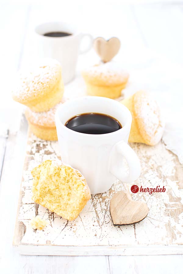 Dieses Foto zum Eierlikörmuffins Rezept ist von schräg oben aufgenommen. Zu sehen ist eien Tasse Kaffee, davor ein angebissener Muffin und ein Holzherz. Im Hintergrund weitere Muffins mit Eierlikör und noch ein Becher Kaffee.