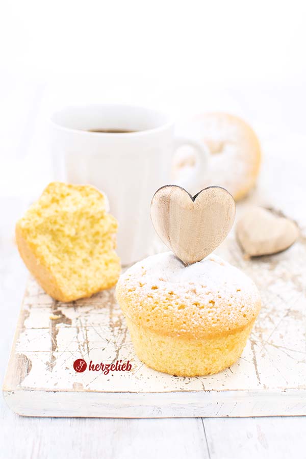 Auf diesem Bild zum Eierlikörmuffins Rezept sieht man im Vordergrund einen Muffin, in dem ein Holzherz steckt Dahinter ist ein angebissener Kuchen vor einer Tasse Kaffee. Dahinter ein weiteres Holzherz und noch einer von den Eierlikör Muffins.