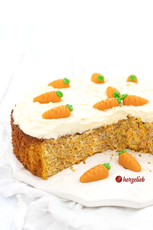 Auf diesem Bild zum Karottenkuchen Rezept ohne Mehl sieht man den angeschnittenen Möhrenkuchen mit einer Frischkäsehaube. Dekoriert mit Marzipan-Möhren. Angerichtet ist der Kuchen auf einem weißen Teller.