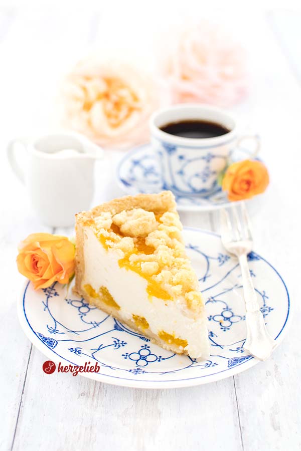 Ein Stück Käsekuchen sieht man auf diesem Bild zum Zitronen-Mandarinen-Käsekuchen Rezept mit einer Prise Liebe. Das Kuchenstück liegt auf einem weißblauem Teller. Hinter dem Kuchen liegt eine lachsfarbene Rose, auf dem Tellerrand liegt eine Kuchengabel aus Silber. Weiter im Hintergrund ein Milchkännchen, eine Tasse Kaffee und Blüten.