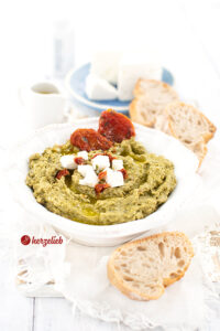 Rezept Grünkohlcreme - Dip in eienr weißen Schale mit Brot, Feta-Würfeln und getrockneten Tomaten dekoriert.
