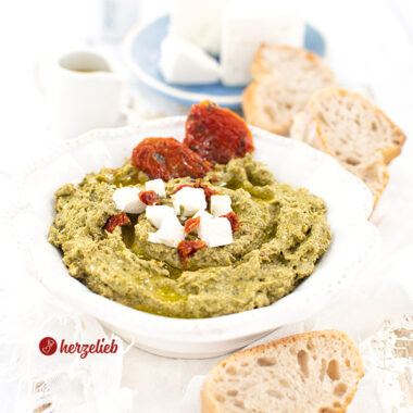 Rezept Grünkohlcreme - Dip in eienr weißen Schale mit Brot, Feta-Würfeln und getrockneten Tomaten dekoriert.