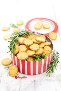 Rezept Rosmarin-Parmesan-Plätzchen. Käsekekse in einer rotweiß-gestreiften dose. Dekoriert mit Rosmarin. Im Hintergrund der Deckel der Dose