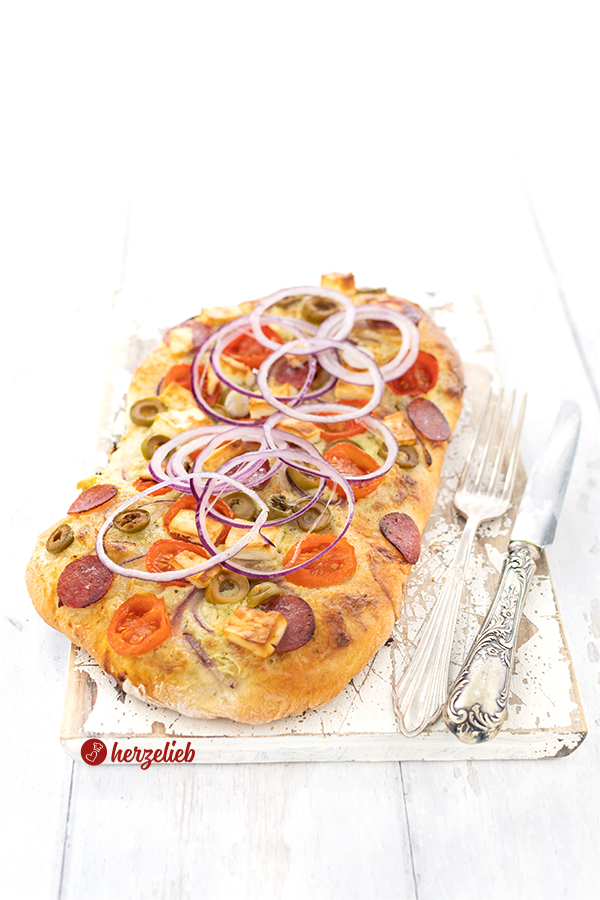 Auf diesem Bild zum Kartoffelpizza Rezept ist eine Salamipizza zu sehen. Belegt mit Tomaten, grüne Oliven und roten Zwiebelringen. Seitlich liegen Messer und Gabel. 
