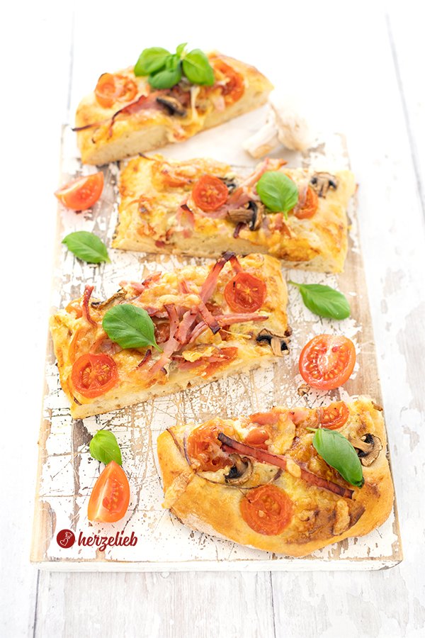Auf diesem Bild zur Kartoffelpizza ist eine aufgeschnittene Pizza, belegt mit Tomaten, Schinken, Pilzen und einer Deko aus Basilikum zu sehen. Zwischen den Stücke sind geviertelte und halbierte, kleine Tomaten und Basilikumblättchen zu sehen.