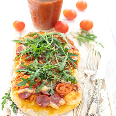 Auf diesem Bild zur Kartoffelpizza ist ein Stück Pizza mit Tomaten, Salami, ;äse und Rucola auf dem knusprigen Pizzaboden zu sehe. Seitlich Messer und Gabel, im Hintergrund selbstgemachte Pizzasauce, Tomaten und Rucola