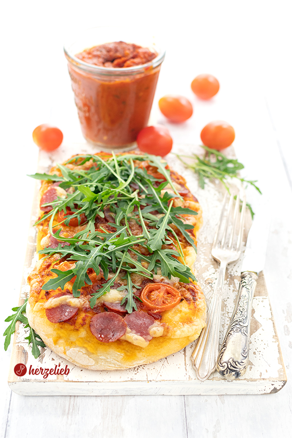Auf diesem Bild zur Kartoffelpizza ist ein Stück Pizza mit Tomaten, Salami, Käse und Rucola auf dem knusprigen Pizzaboden zu sehe. Seitlich Messer und Gabel, im Hintergrund selbstgemachte Pizzasauce, Tomaten und Rucola