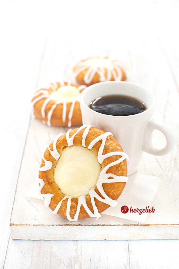Auf diesem Bild zum Pudding-Donut sieht man einen Kuchen an einem Becher Kaffee gelehnt. Der goldbraun gebackene Donut ist mit Zuckerguss verziert und in der Mitte ist Vanillepudding