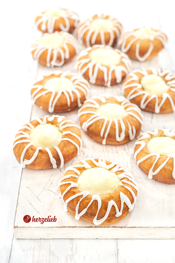 Auf diesem Bild zum Pudding-Donuts Rezept ist ein Brett mit 12 Kuchen zu sehen. Goldbraun gebacken, mit Zuckerguss verziert und mit Vanillepudding gefüllt. 