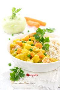 Dieses Bild zum Kohlrabi Rezept zeigt die Kohlrabi-Currypfanne von vorne fotografiert in einer weißen Schüssel. Man sieht die Kohlrabiwürfel und die Karottenwürfel, die Erbsen und das Hähnchenfleisch in der gelben, cremige Sauce. Im Hitnergrund Petersilie, ein Kohlrabi und 2 Karotten. Im Vordergrund Erbsen und glatte Petersilie.