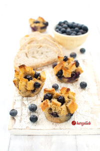Auf diesem Bild zum Arme Ritter muffins Rezept sieht man 3 Muffins mit Blaubeeren, oben knusprig gebacken. Im Hintergrund eine gelbe Schale mit Blaubeeren, 3 Scheiben Brot und ein weiterer Muffin.